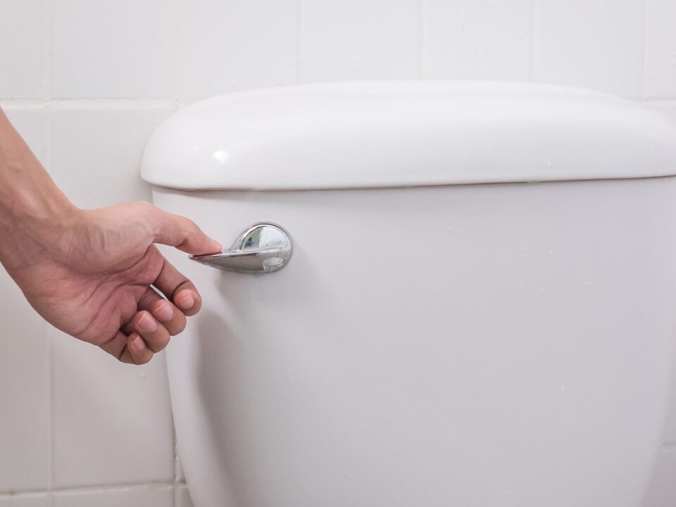 Hand flushing a modern white toilet using a chrome flush handle in a residential bathroom